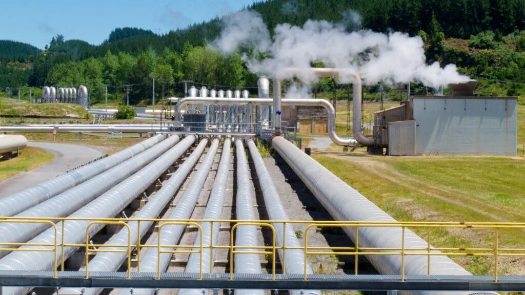 A geothermal power plant with multiple silver pipelines and steam rising against a backdrop of green hills.