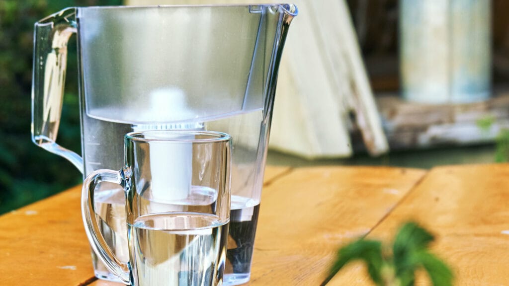 Transparent water pitcher and glass filled with purified well water on a wooden table.