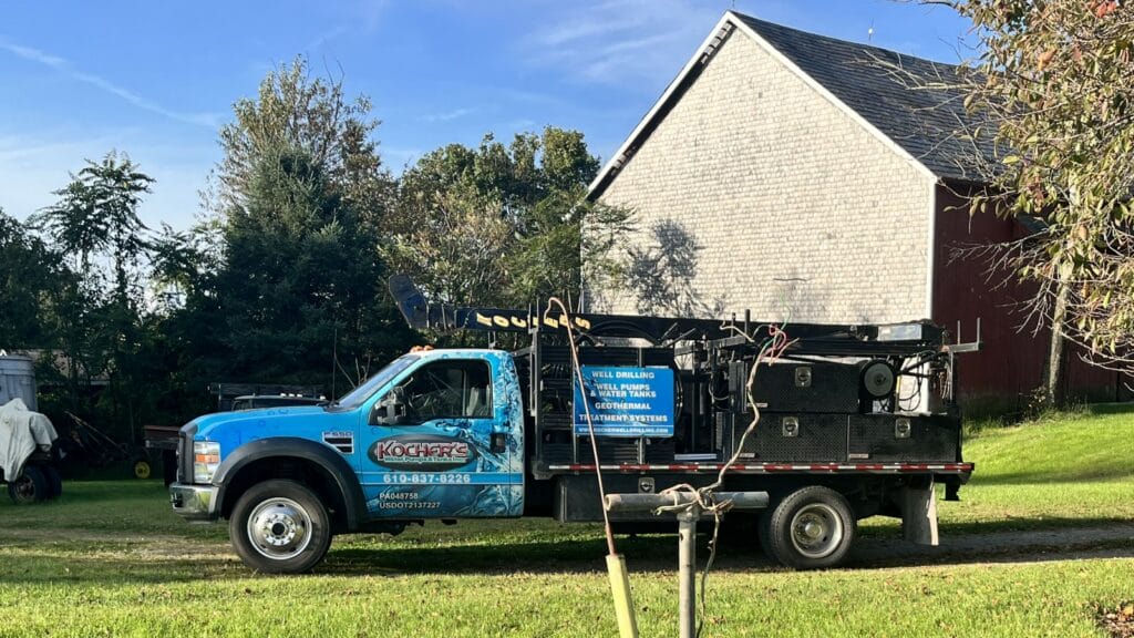 A blue water well drilling truck parked on grass with equipment and signage for well drilling, water pumps, and geothermal treatment systems, in front of a house.